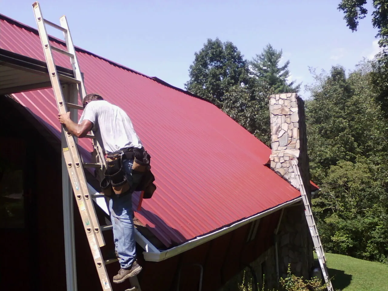 Roofer on a ladder beside a red metal roof and stone chimney — Soffit & Fascia Repair by Roofing Brothers in Atwater